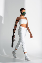 futuristic african american woman in safety mask walking with assault rifle on white background © LIGHTFIELD STUDIOS