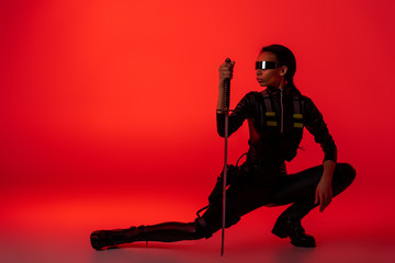 futuristic african american woman in glasses sitting with sword on red background