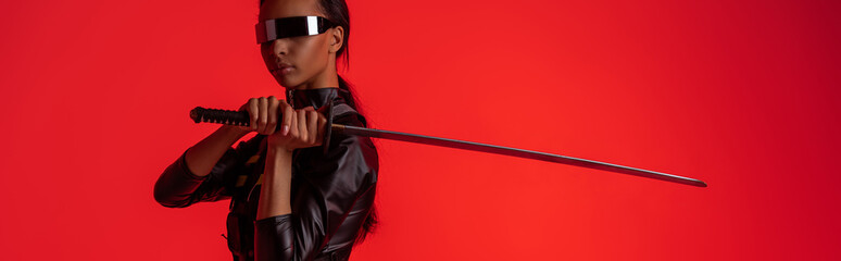 attractive futuristic african american woman in glasses with sword isolated on red, panoramic shot © LIGHTFIELD STUDIOS