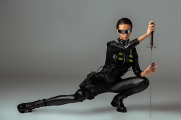 attractive futuristic african american woman in glasses sitting with sword on grey