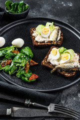 Sandwiches with sardines, egg, cucumber and cream cheese, salad garnish with spinach and dried tomatoes. Black background. Top view