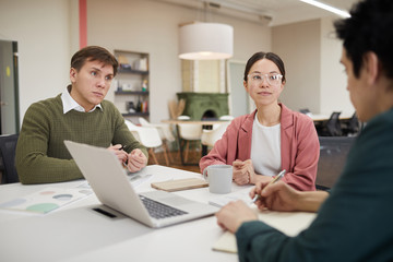 Young business people sitting at the table and listening to their leader during meeting at office