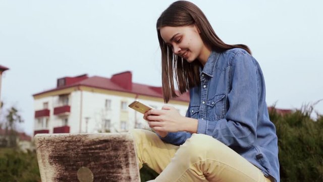 Young laughing woman in casual clothes is recording audio messages and chatting on social network while sitting on stone parapet in front of building