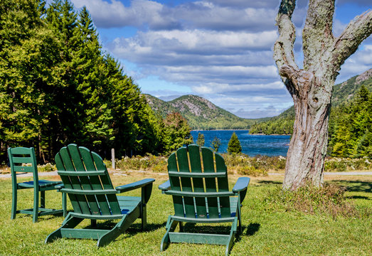 Two Green Adirondack Chairs In A Park Overlooking A Blue Lake