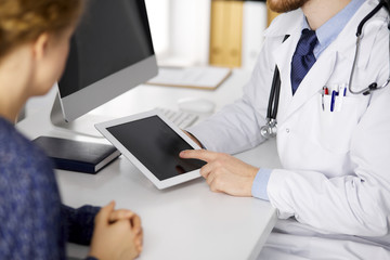 Unknown red-bearded doctor and patient woman discussing current health examination while sitting and using tablet computer in clinic, close-up. Medicine concept