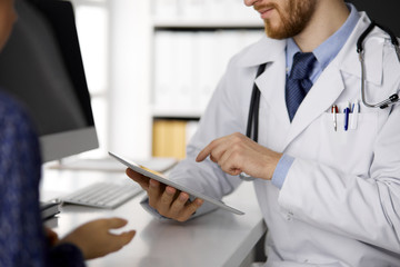 Fototapeta premium Unknown red-bearded doctor and patient woman discussing current health examination while sitting and using tablet computer in clinic, close-up. Medicine concept