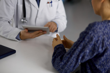 Unknown red-bearded doctor and patient woman discussing current health examination while sitting and using tablet computer in clinic, close-up. Medicine concept