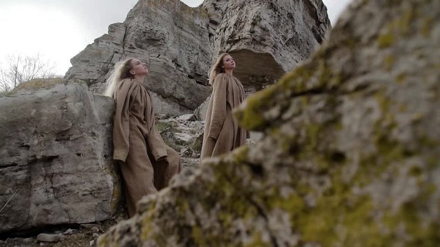 two young beautiful blonde girls in long dresses are standing on the stone mountains, their hair and clothes are developing in the wind, they are sad