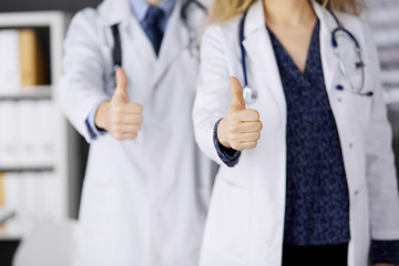 Two doctors standing with thumbs up in hospital office. Medical help, countering viral infection and medicine concept