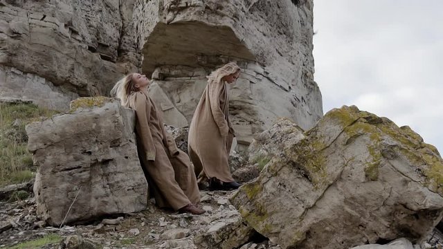 two young beautiful blonde girls in long dresses are standing on the stone mountains and looking at the sky, their hair and clothes are developing in the wind, the ancient world