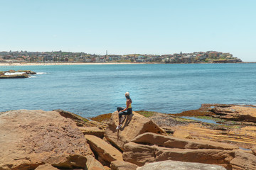 Woman at Bondi Beach. Girl standing on rocks in work out gear looking at view of the ocean, sun, sea and sand scene while on vacation. Holiday, tropical, explore, fitness. Sydney, Australia