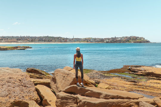 Woman at Bondi Beach. Girl standing on rocks in work out gear looking at view of the ocean, sun, sea and sand scene while on vacation. Holiday, tropical, explore, fitness. Sydney, Australia
