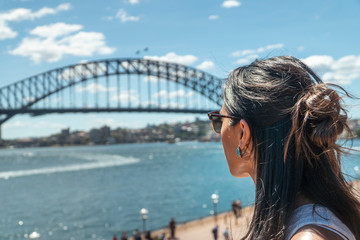 Woman at Iconic Sydney Harbor Bridge over river water. Cityscape with buildings in CBD area. Tourist attraction in Australia. People at busy landmark. Black steel construction.