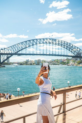 Woman taking selfie with mobile phone at iconic Sydney Harbor Bridge. Cityscape, water, with...