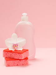 Bottle of dishwashing liquid, sponges and an orchid flower on a pink background.