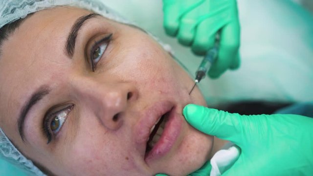 CU: cosmetologist in protective gloves does anti-aging facial filler subcutaneous injection to woman client in modern beauty clinic close view