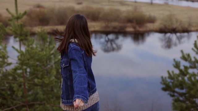 Cheerful young woman in casual clothes is spinning around and enjoying picturesque landscape while walking in nature on hill along river