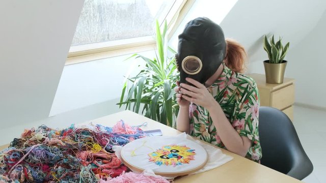 girl wears gas mask at home during coronavirus pandemic COVID-19, sews with needle. embroidery hoop, fabric with colorful image. Stay home, quarantine leisure funny concept. Needlework, handicraft