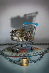 Small shopping cart with Money in Euro coins and banknotes. Dark and ominous setting, spooky. Chain with safety lock for protection against thieves and criminals.
