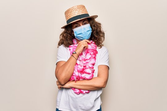 Middle age tourist woman wearing coronavirus protection mask and summer hat on vacation smiling looking confident at the camera with crossed arms and hand on chin. Thinking positive.