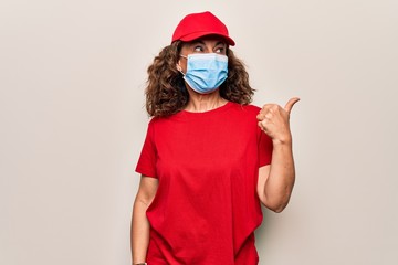 Fototapeta na wymiar Middle age delivery woman wearing red cap and coronavirus protection mask pointing thumb up to the side smiling happy with open mouth