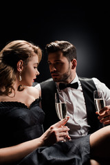 sexy woman and elegant man holding glasses of champagne and looking at each other on black background