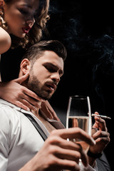 sexy girl touching neck of handsome, confident man holding champagne glass and smoking isolated on black