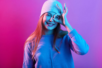 Closeup portrait of smiling teenager girl posing against pink neon wall and covering her eye with ok sign, woman wearing sweater and hat, expresses positive emotions, looks satisfied. People concept - obrazy, fototapety, plakaty