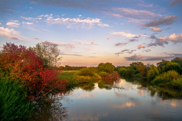 Obraz premium Sunset over the river. Clouds are reflected in the river at sunset, along the banks of which trees with reddening foliage