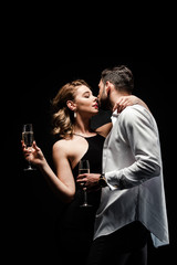 handsome man in unbuttoned shirt and elegant, sexy woman holding champagne glasses and kissing isolated on black