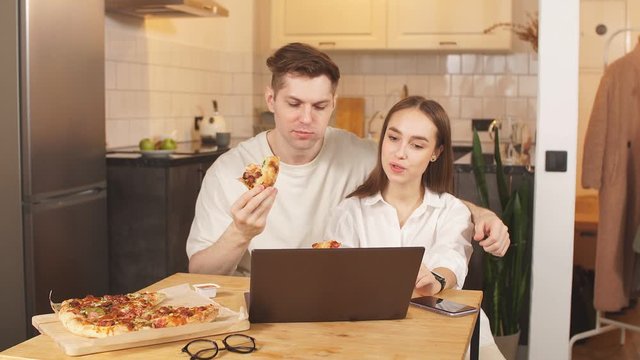 Young happy couple eat pizza and watch film at home. Married couple look at screen of laptop, enjoy free time at home together. Relationships, food concept