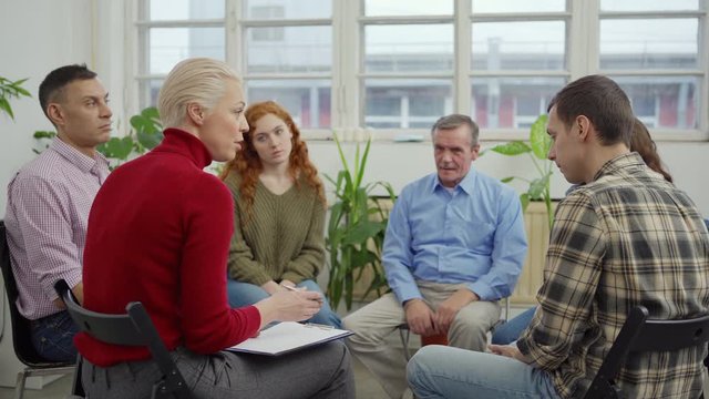 Tracking wide shot of middle aged female therapist with clipboard talking to patients nodding heads sitting in circle during group therapy session