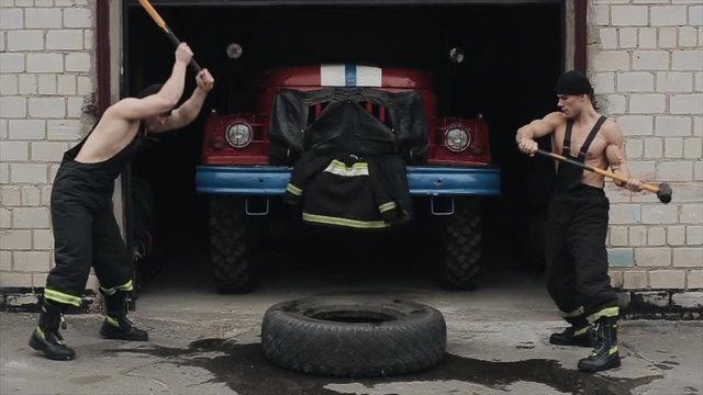 Two strong muscular fireman hit with a hammer on a large tire against the background of a fire engine in a garage. Firemans doing exercises and workout