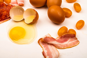 Broken chicken egg, slices of sausage and bacon, tomatoes and chicken eggs scattered on a white background. Close up.