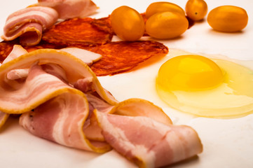 A broken chicken egg without a shell, slices of sausage and bacon and tomatoes on a white background. Close up.