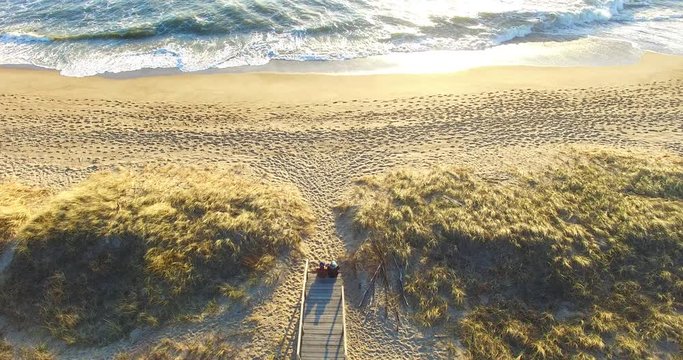 Aerial dinamic view from a top that starts from a couple sitting on the steps that lead to the beach, that is full of human traces in the sand, and goes to the rough green sea with big foamy waves