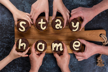 Concept flat lay Stay Home. Many hands of different ages of people reach out and hold chocolate muffins, with cream letters. Eight cupcakes make up the two words. Big family on self-isolation. - Powered by Adobe