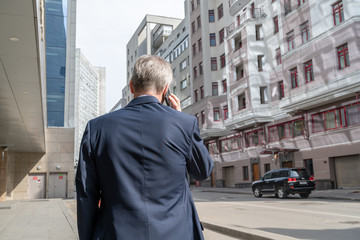 Alone middle-aged gray-haired businessman in a blue suit calling by phone standing on the empty city street.