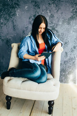 A cute long-haired girl in a blue shirt and dark blue jeans is reading an e-book, perched with her feet on a chair - 339912759