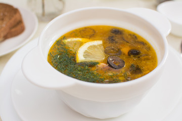 Traditional Russian meat soup solyanka with sausages, lemon and olives in a white ceramic bowl.