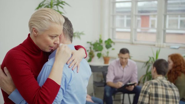 Tracking medium shot of depressed middle aged woman and senior man hugging to console each other during group therapy session, therapist and other patients sitting in circle in background