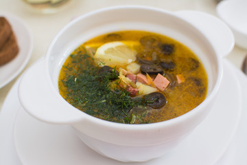 Traditional Russian meat soup solyanka with sausages, lemon and olives in a white ceramic bowl.