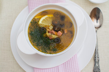 Traditional Russian meat soup solyanka with sausages, lemon and olives in a white ceramic bowl.