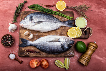 Two Dorado fish lie on a wooden chopping Board on beautiful pink concrete. Next are: lime, lemon, pepper, tomato, garlic, spices, salt and herbs.