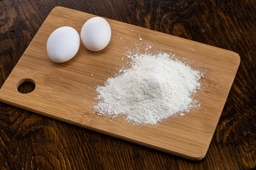 two white eggs on a wooden board and next to a handful of flour