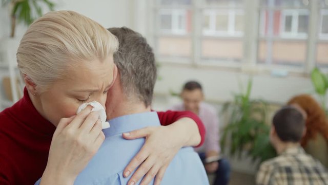 Closeup of depressed middle aged woman crying and hugging senior man at group therapy session, therapist and other patients sitting in circle in background