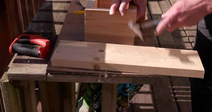 A man building a bird-box by hammering nails into wood.