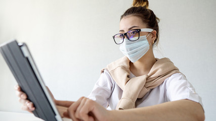 serious girl in a surgical mask works at home on a tablet. girl in hipster glasses works at home, self-isolation. Coronavirus outbreak or Covid-19, Concept of Covid-19 quarantine
