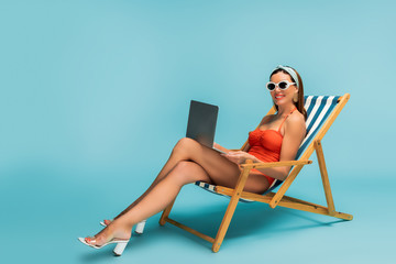 Freelancer with crossed legs and laptop smiling on deckchair on blue