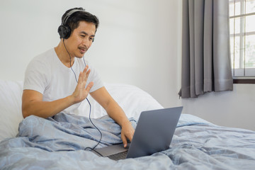 Man with headphones sitting in bed and waving hand having video call on laptop coronavirus quarantine.